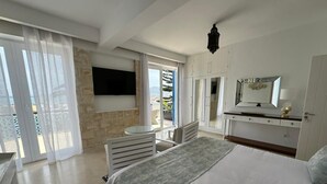 Un dormitorio con cama, televisor montado en la pared, escritorio y una silla. Hay un balcón con vista al mar.