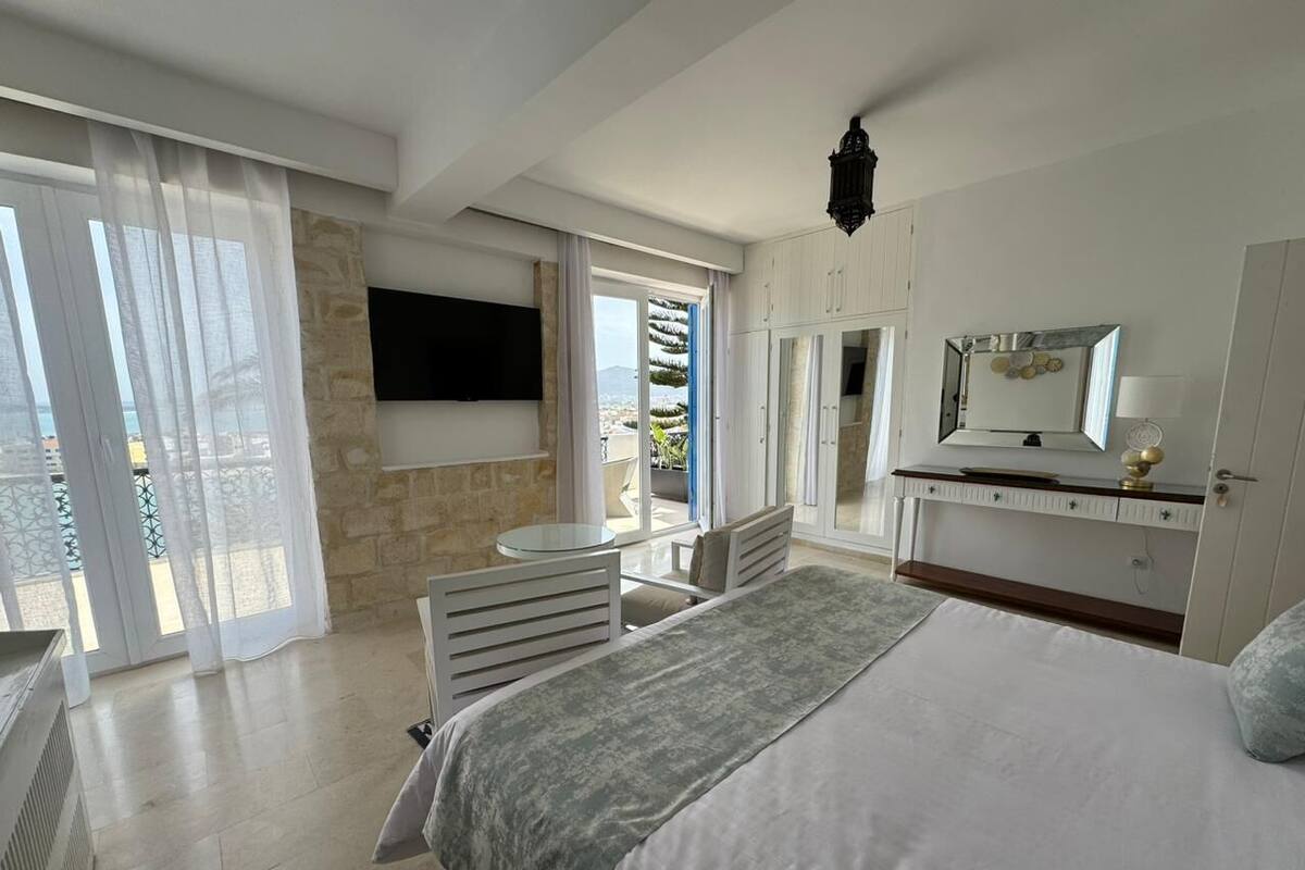 Ein Schlafzimmer mit Bett, Wand-Fernseher, Schreibtisch und Stuhl. Ein Balkon bietet Blick aufs Meer.