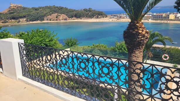 Un balcón con piscina, con vista a una zona costera que incluye una playa y una colina con una estructura similar a un castillo.