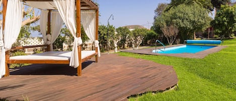A wooden deck with a four-poster bed and a swimming pool in the background.