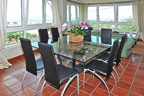 A dining area with a glass table surrounded by chairs, a woven centerpiece, and large windows offering a view of the sea.
