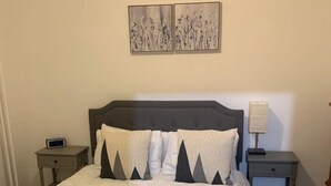 A neatly made bed with a grey and white duvet, two pillows with geometric patterns, a bedside table with a lamp, and a wall clock.