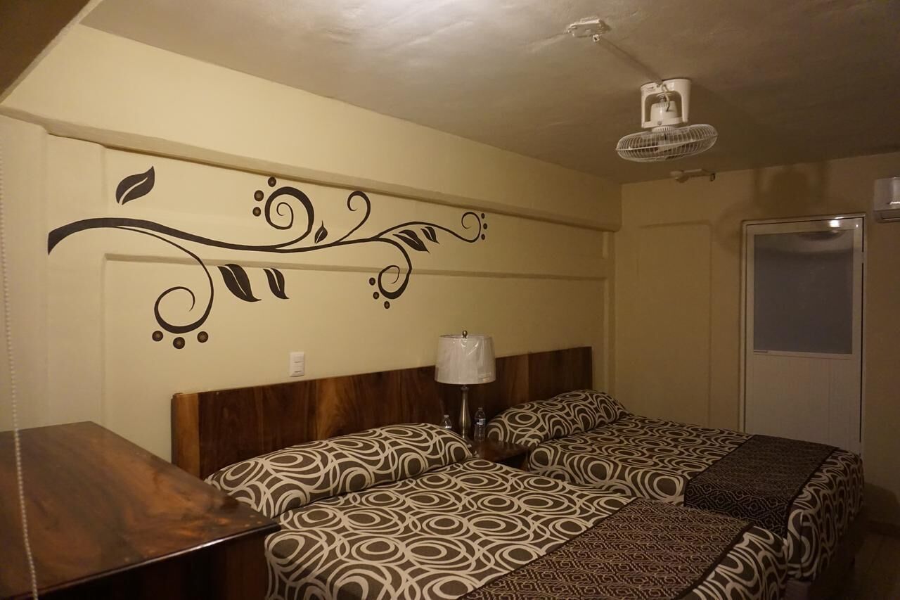 A hotel room with two beds, a wooden headboard, a wall decoration, a bedside table with a lamp, and a ceiling fan.