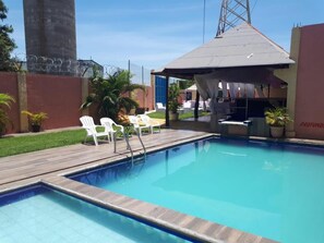Una piscina con muelle de madera, sillas blancas y una zona de estar cubierta.