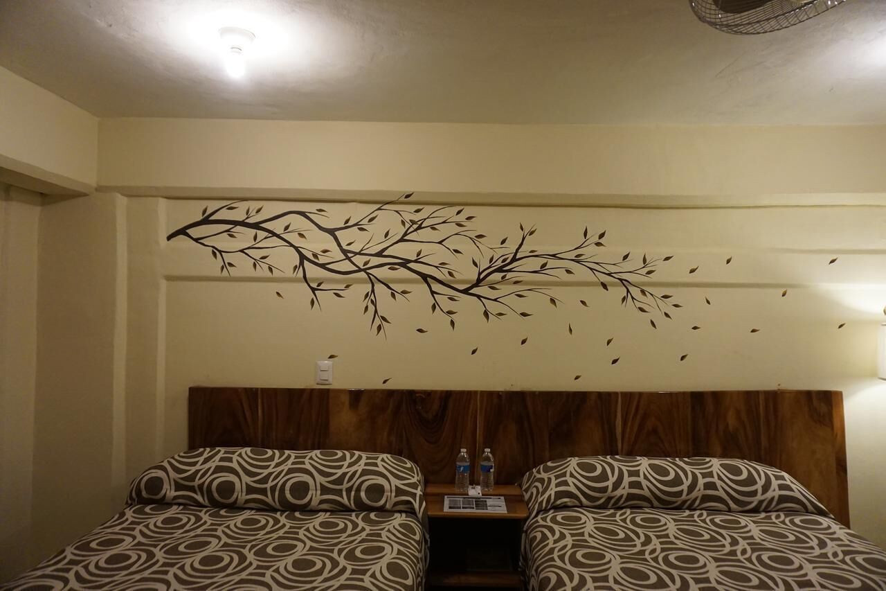 Habitación de hotel con dos camas estampadas, cabeceros de madera, una decoración de rama montada en la pared y una mesita de noche con botellas de agua.
