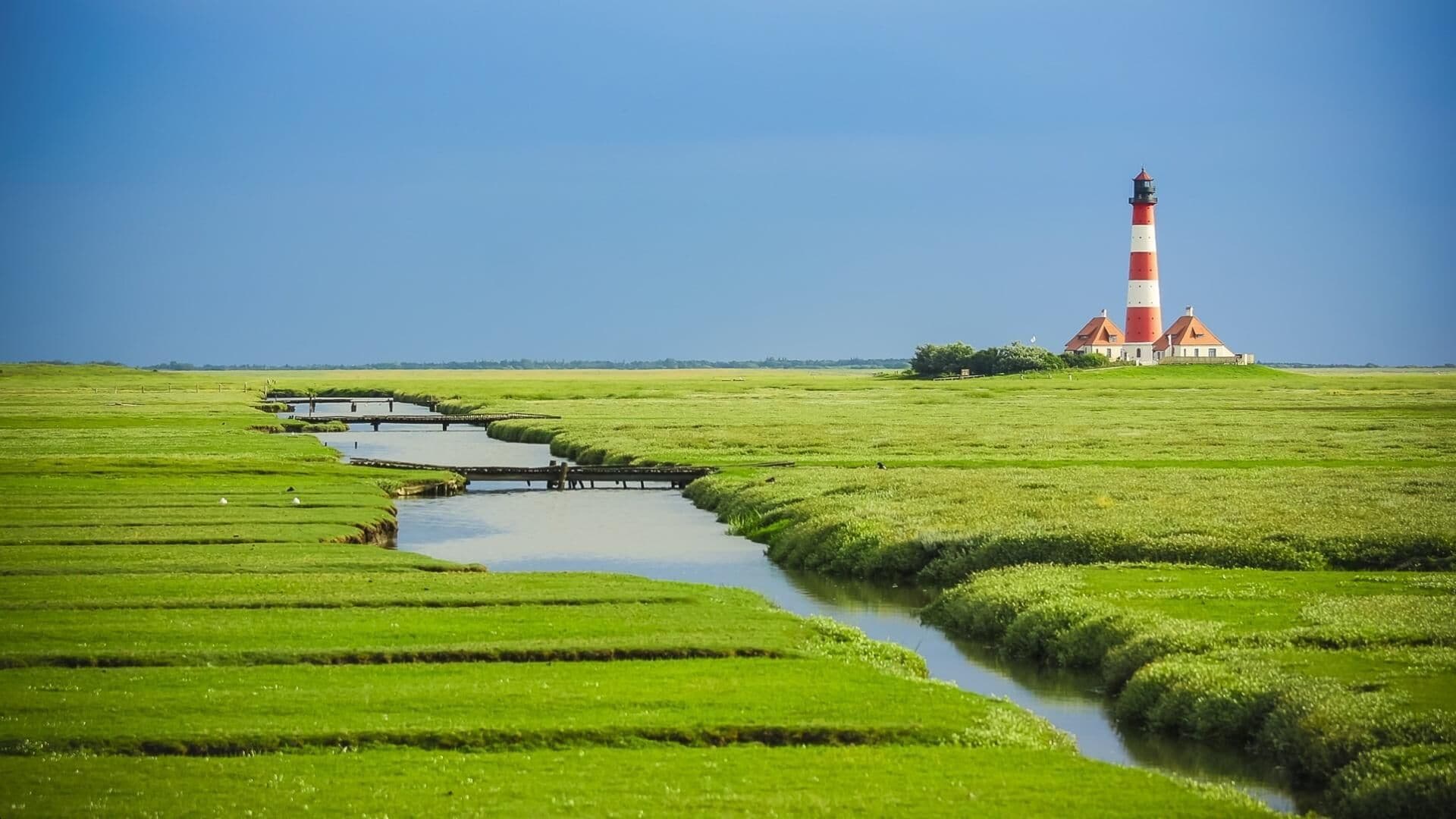 A lighthouse with a red and white striped tower, a red roof, and a white building with a red roof in the background. There is a small water channel with a wooden bridge crossing it.