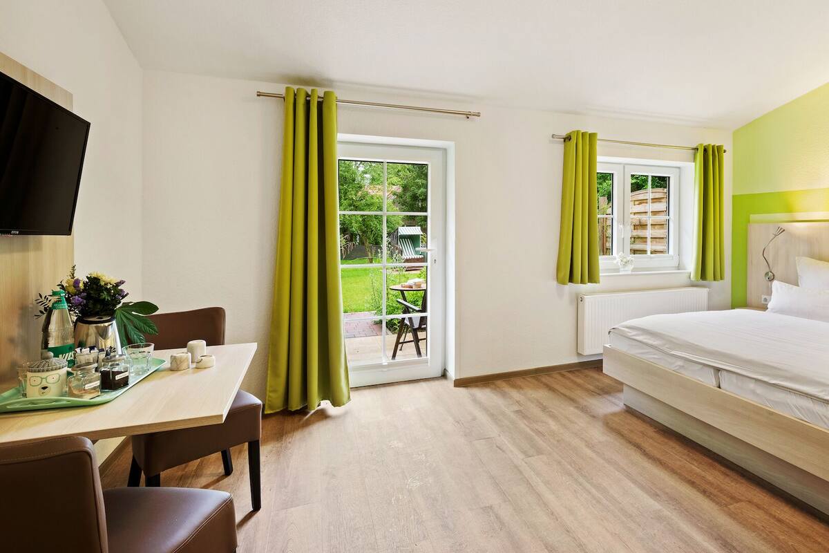 A hotel room with a bed, a desk with a chair, a television, and a view of the garden through the window.