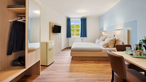 Et lille hotelværelse med seng, skrivebord med stol, garderobeskab, fjernsyn og et vindue med gardiner.