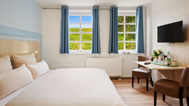 Et hotelværelse med en stor seng, et skrivebord med stol, et fjernsyn og to vinduer med gardiner.