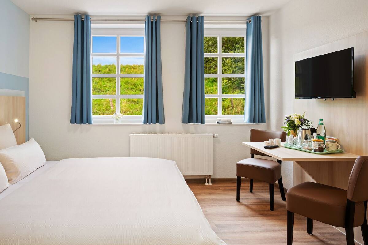 A hotel room with a large bed, a desk with a chair, a TV, and two windows with curtains.