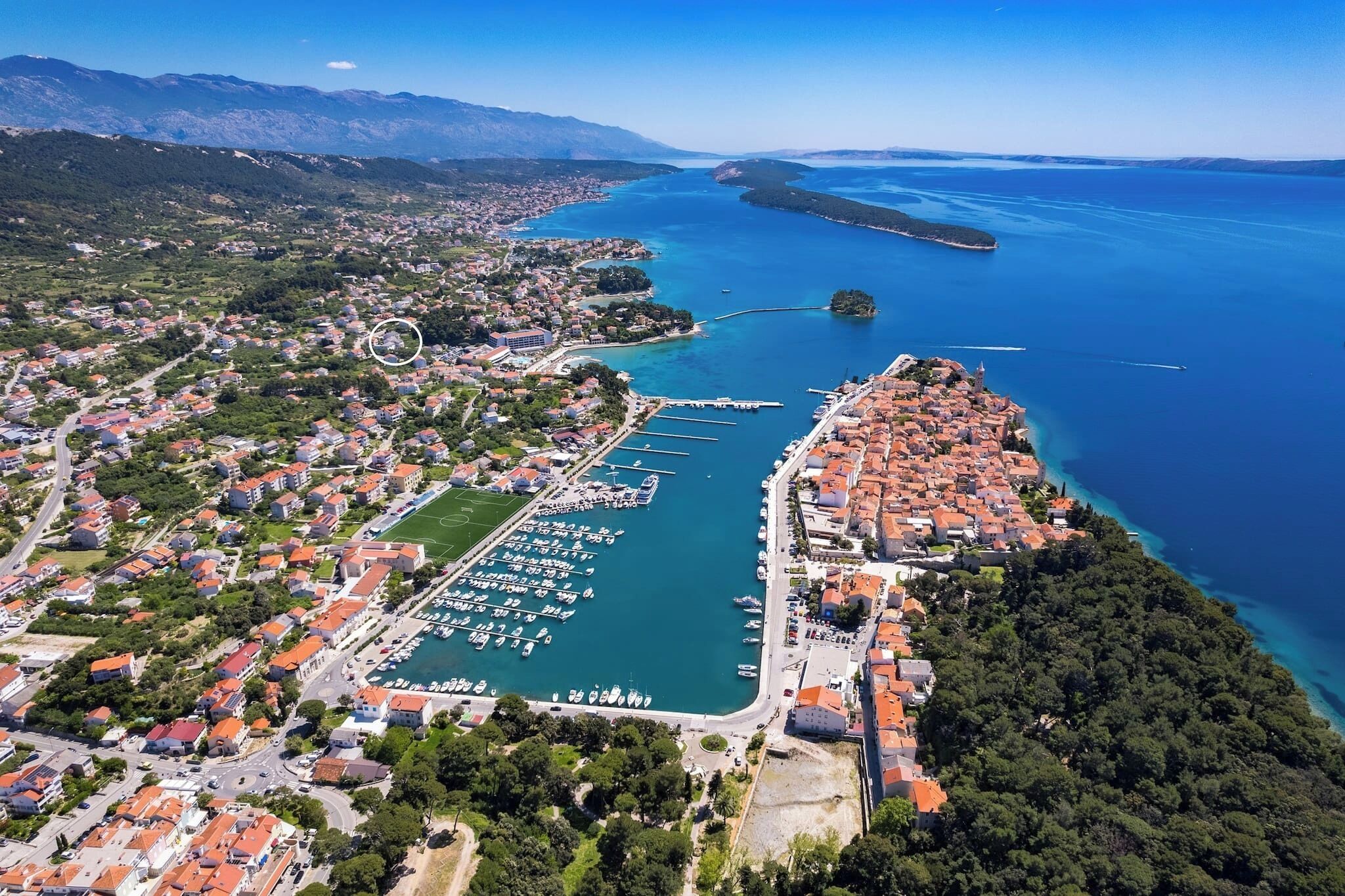 An aerial view of a coastal town with a marina, residential areas, and a mountainous backdrop.