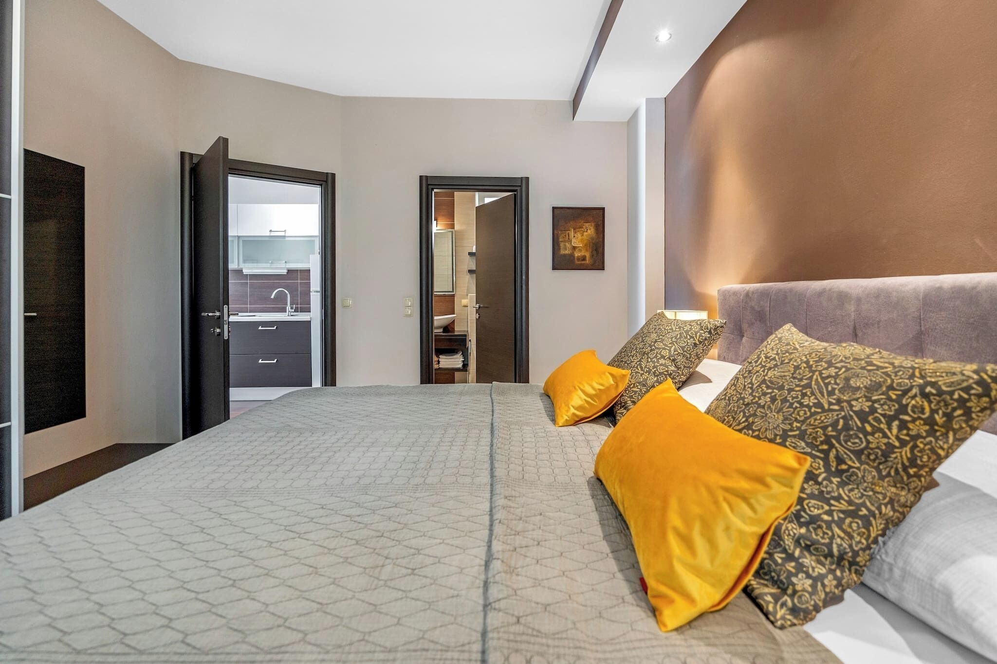 A modern hotel room with a large bed, a sofa, and a small table. There is a door leading to a bathroom and a hallway.