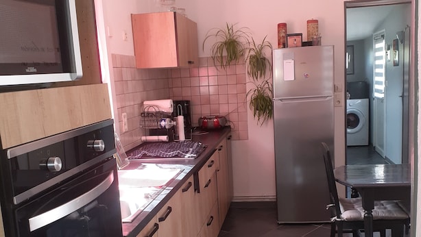 Moderni keittiö, jossa on puukehikoiset kaapit, sisäänrakennettu uuni, mikroaaltouuni ja jääkaappi. Työtasolla on ruukkukasveja ja keittiössä on ruokailutila, jossa on pöytä ja tuolit.