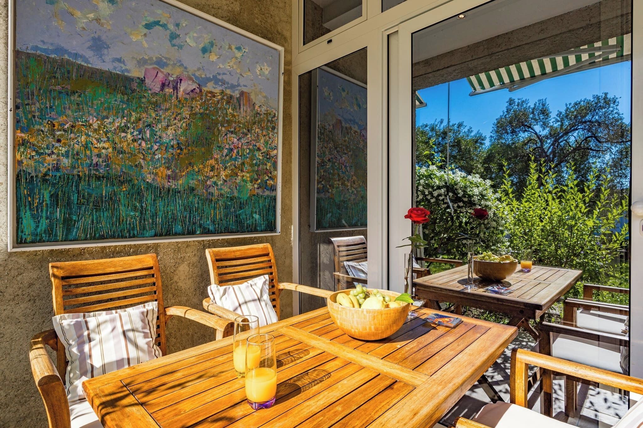 An outdoor dining area with wooden furniture, a table with a bowl of fruit and drinks, and a large painting on the wall.