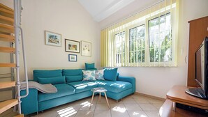 A living room with a blue sofa, a small table, and a television. There are framed pictures on the wall and a window with yellow blinds. - Magical Garden with Shared Jacuzzi - Blanka C2 (Banjol)
