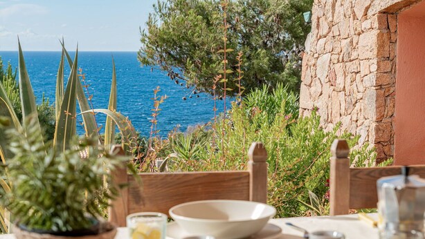 Une table dressée pour le dîner, avec vue sur la mer, entourée de verdure et bordée d’un mur de pierres.