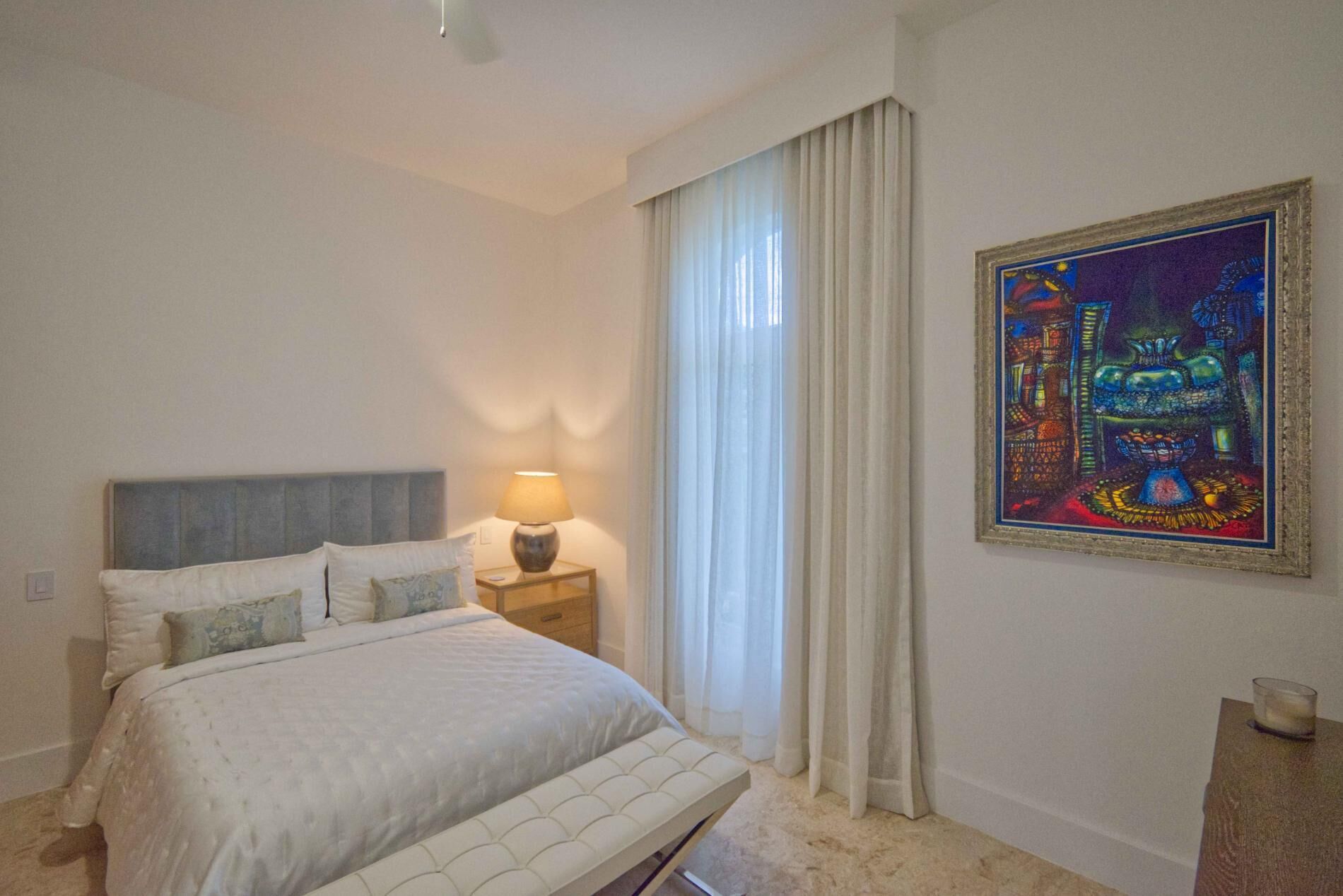 A hotel room with a bed, a nightstand with a lamp, a window with curtains, and a framed painting on the wall.