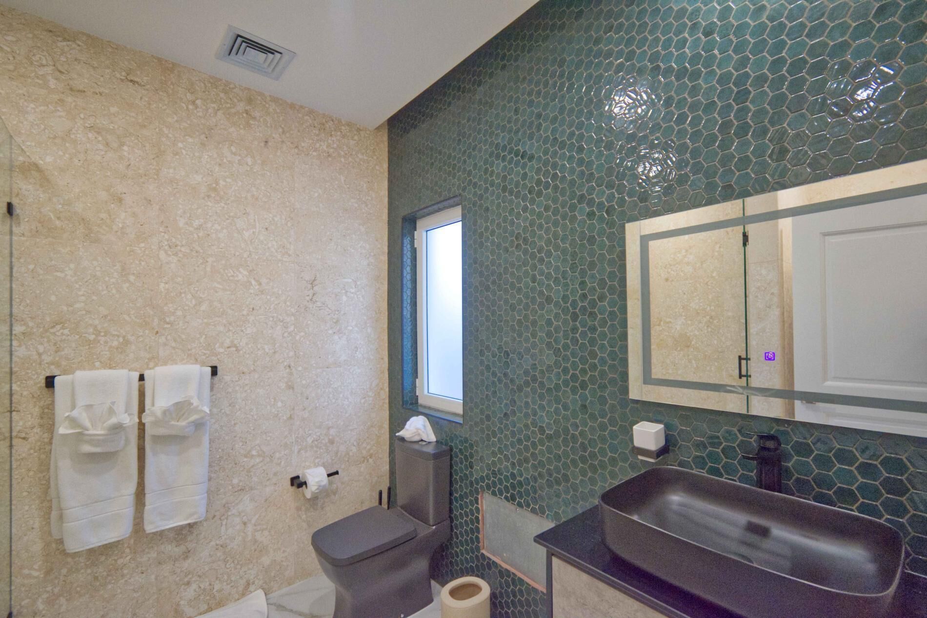 Um banheiro com parede bege texturizada, painel de mosaico verde atrás da pia, pia preta, um espelho e vaso sanitário.