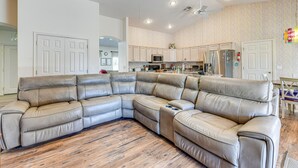 A spacious living room with a large L-shaped sectional sofa, a kitchen area with white cabinets, and a ceiling fan.