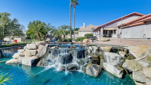 A clear blue pool with a waterfall feature surrounded by rocks, a bridge, and a building with a red roof.