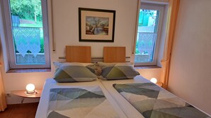 A neatly made bed with striped bedding, two bedside lamps, a framed painting on the wall, and a window with a view of greenery.