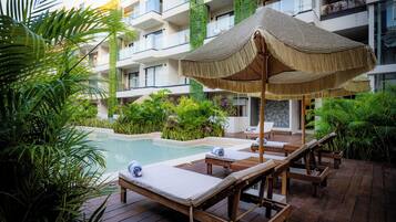 A poolside area with lounge chairs and a thatched umbrella, surrounded by lush greenery and a multi-story building with balconies.