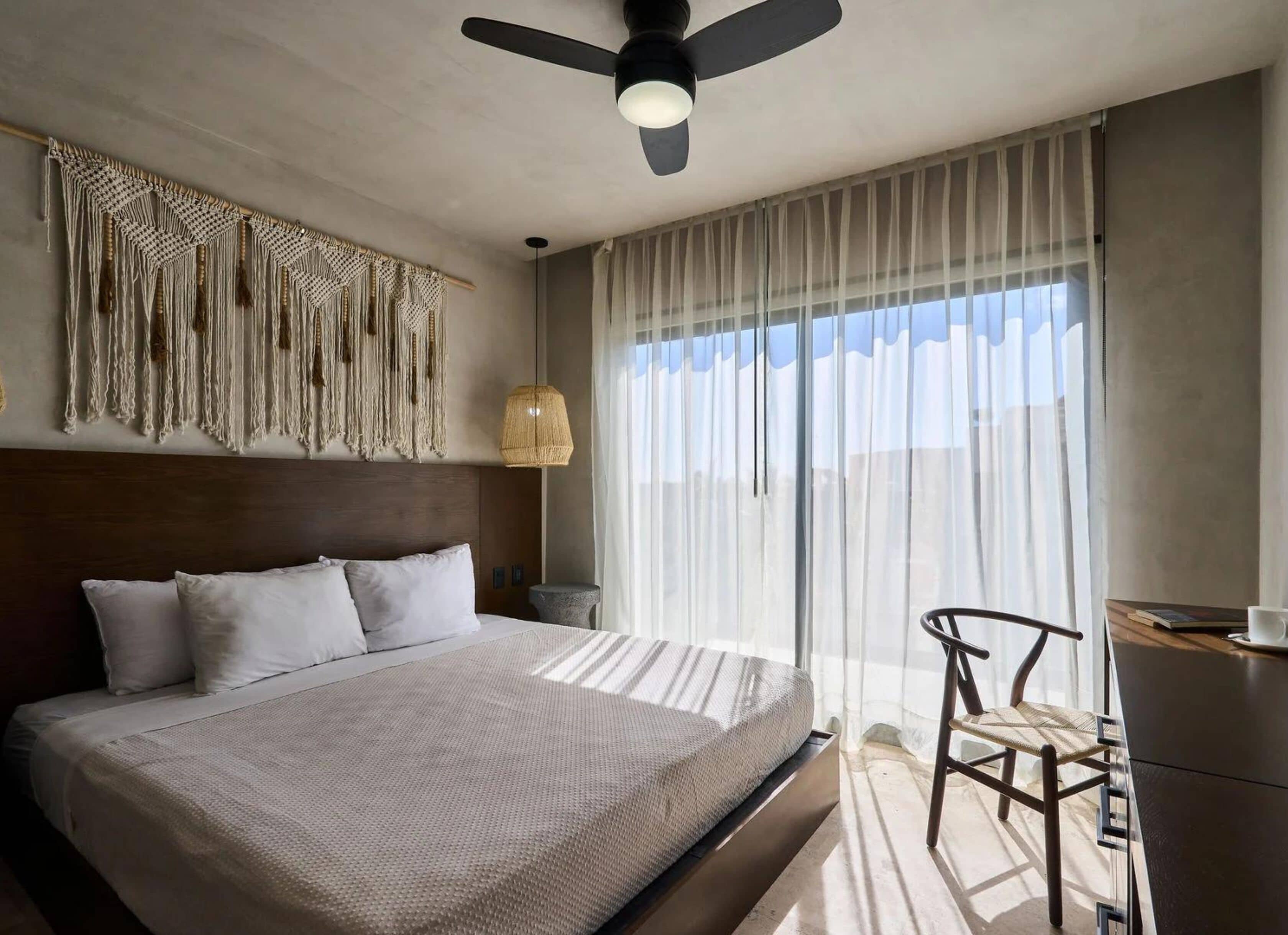 Una habitación de hotel moderna con una cama grande, un ventilador de techo, un escritorio con una silla y un ventanal con cortinas traslúcidas.