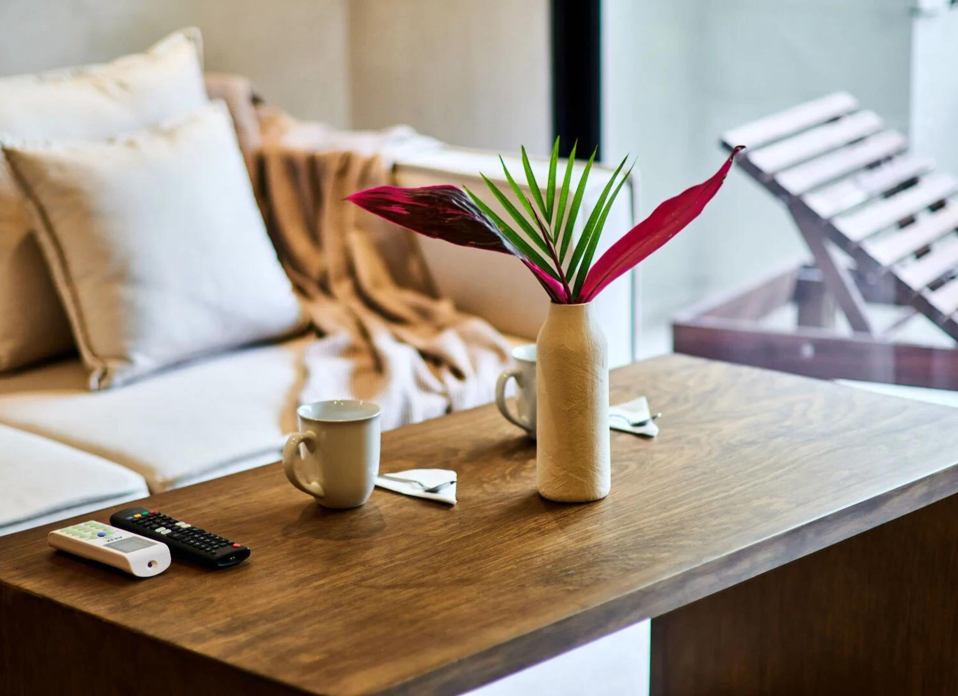 Una mesa de madera con un jarrón beige que contiene hojas verdes y rojas, una taza blanca y dos controles remotos.