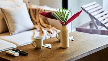 Una mesa de madera con un jarrón beige que contiene hojas verdes y rojas, una taza blanca y dos controles remotos.