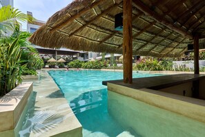 A swimming pool with a thatched roof structure, surrounded by lush tropical plants and a modern building in the background. - Bakal Residences by Ruha (Tulum)