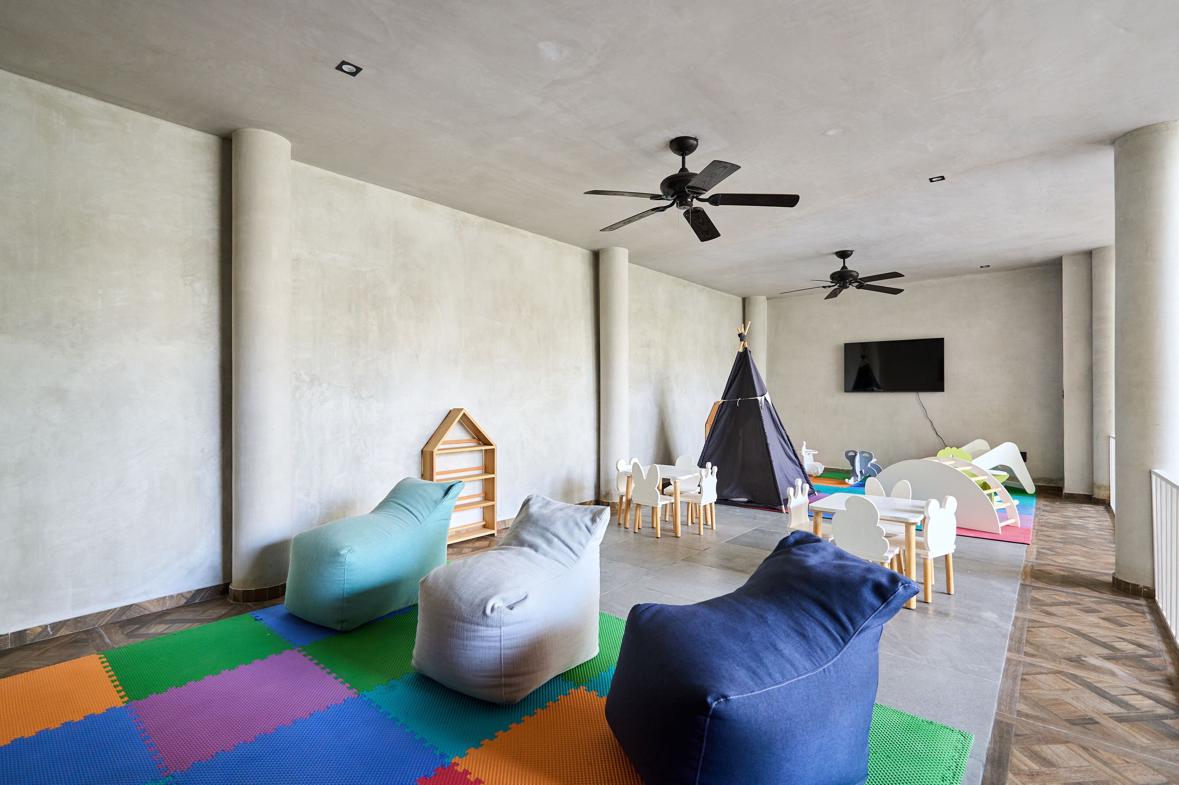 A spacious room with a high ceiling, two ceiling fans, a large window, and a play area with colorful mats and furniture.