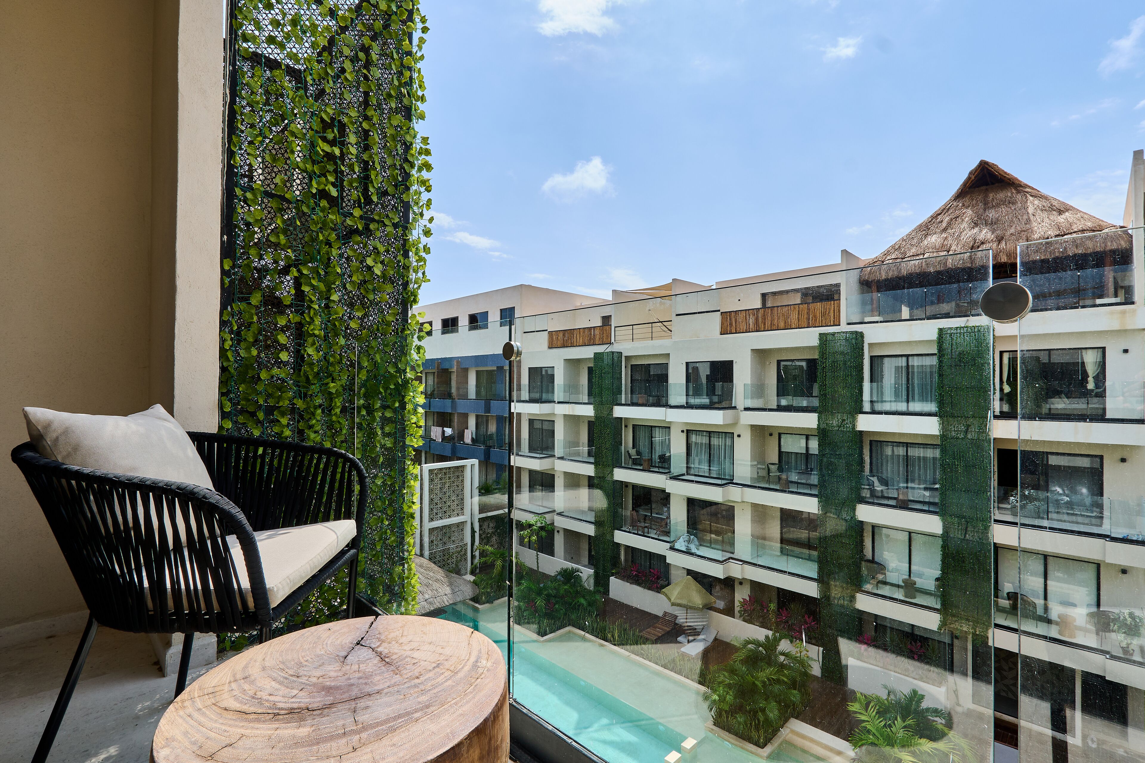 Un balcon agrémenté d’une chaise en osier et d’une table ronde en bois, d’où l’on peut admirer une piscine et un bâtiment doté de nombreux balcons et d’un toit de chaume.