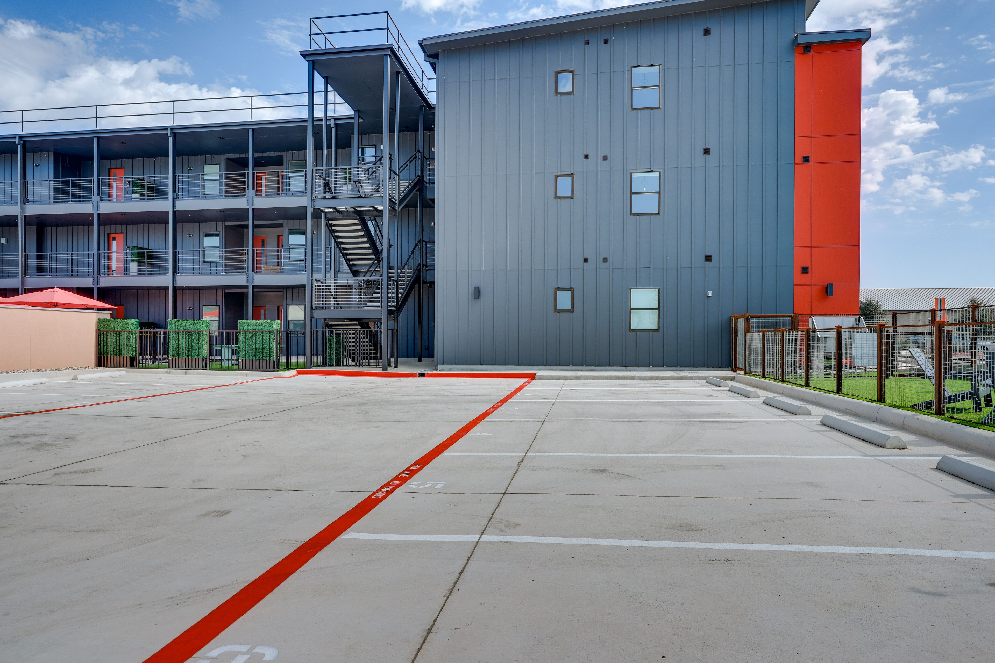 Ein mehrgeschossiges Gebäude mit roter und grauer Fassade, einem Parkplatz mit markierten Stellplätzen und einer Treppe, die zu einer oberen Ebene führt.