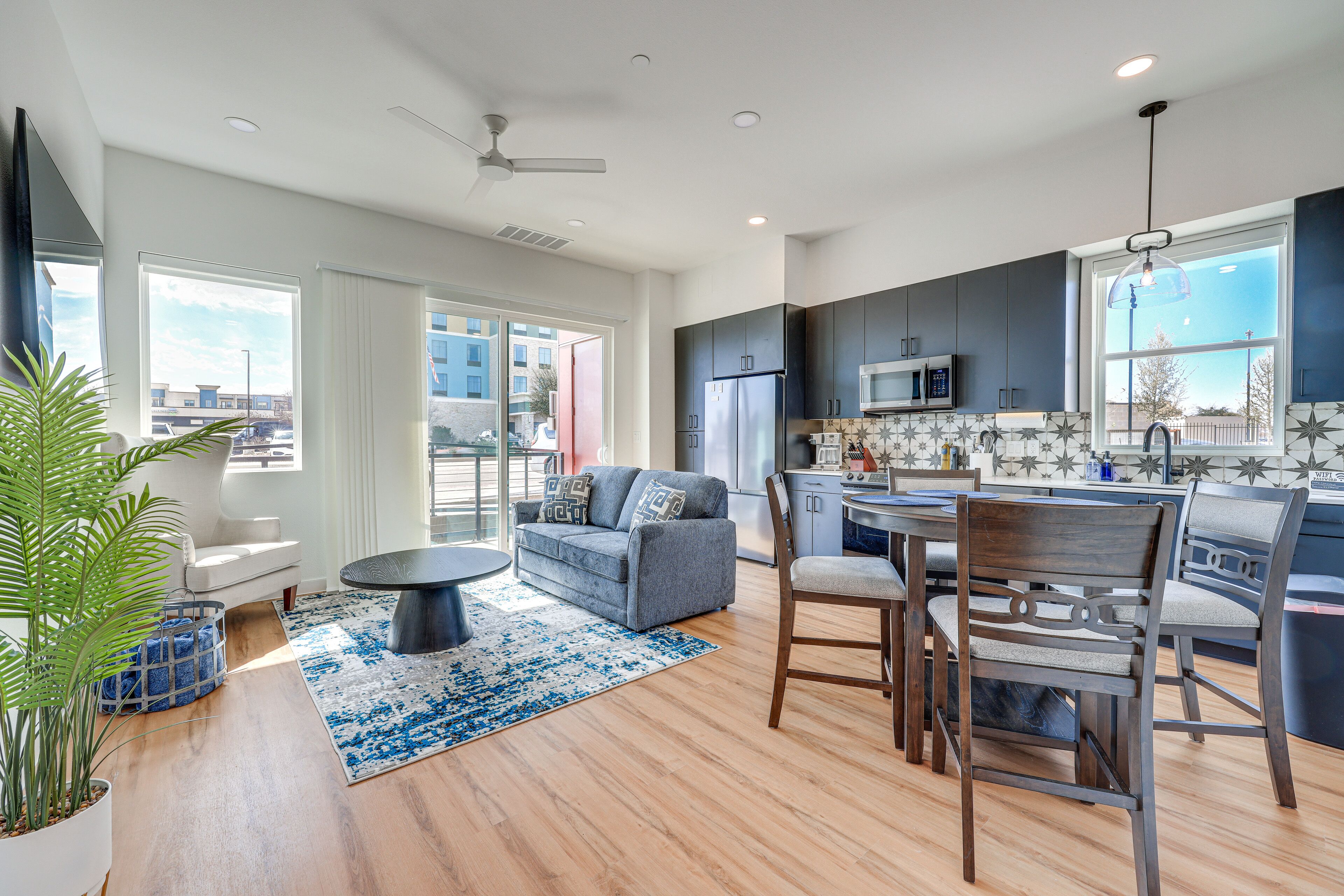 A modern kitchen with dark cabinets, a microwave, and a refrigerator. There is a dining area with a table and chairs, and a large window providing a view of the outside. - Pet-Friendly New Braunfels Condo w/ Shared Pool! (New Braunfels)