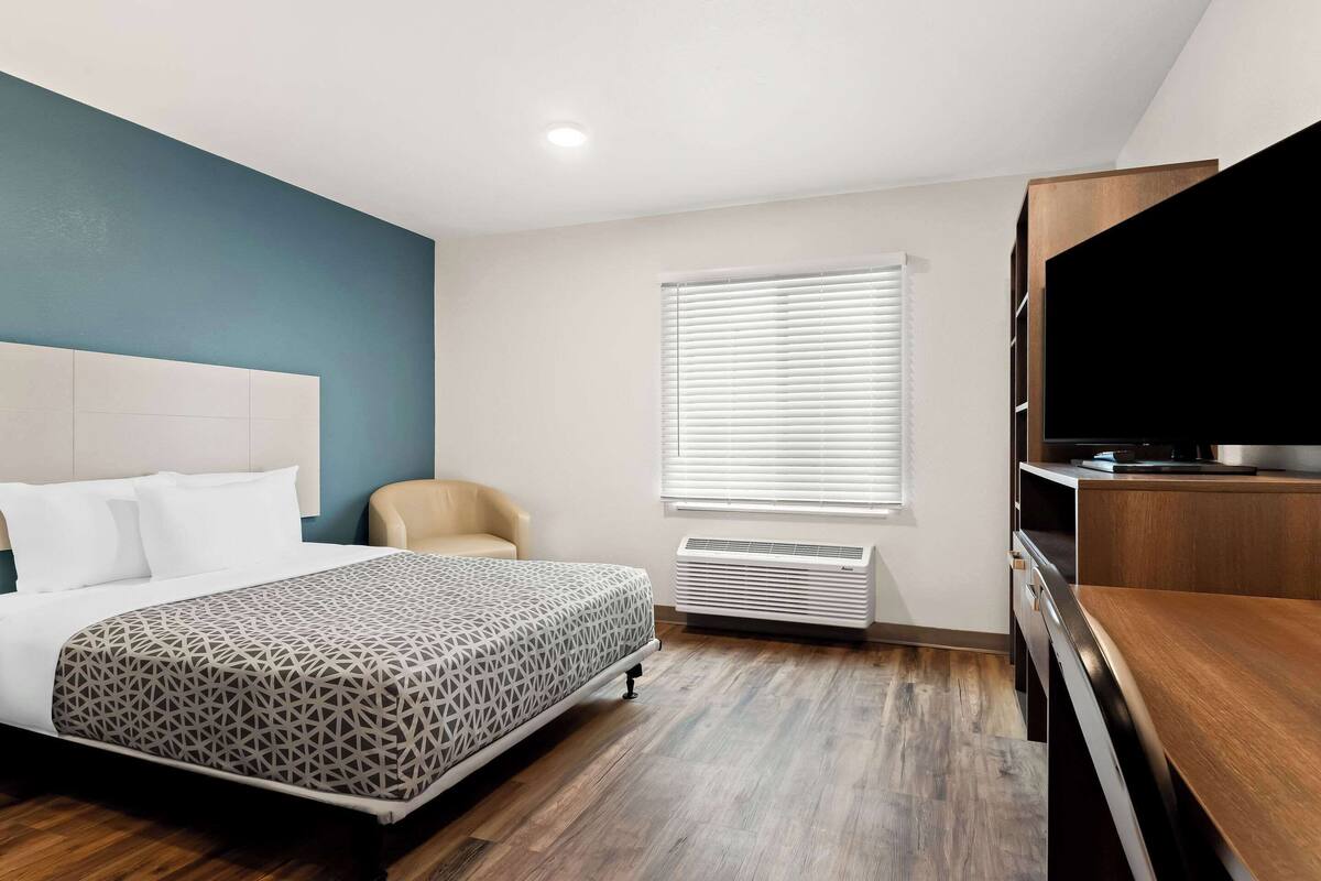 A hotel room with a bed, a chair, a TV, and an air conditioning unit.