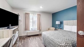 Habitación de hotel con cama, escritorio con computadora y control remoto, una silla, ventana con cortinas y mesita de noche con lámpara.