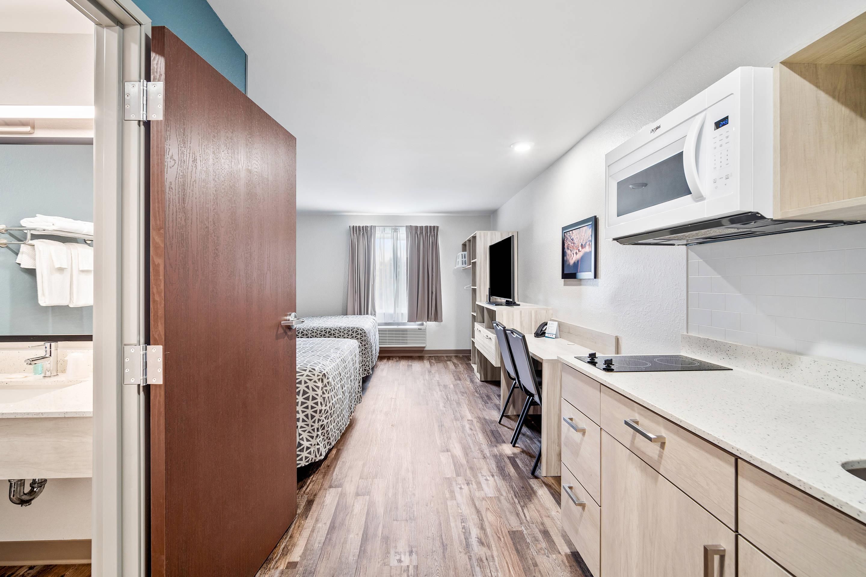 Habitación de hotel compacta con kitchenette, una cama con ropa de cama estampada, un escritorio con silla y un baño con lavamanos y toallero.