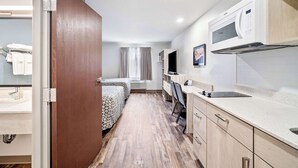 Habitación de hotel compacta con kitchenette, una cama con ropa de cama estampada, un escritorio con silla y un ventanal con cortinas.
