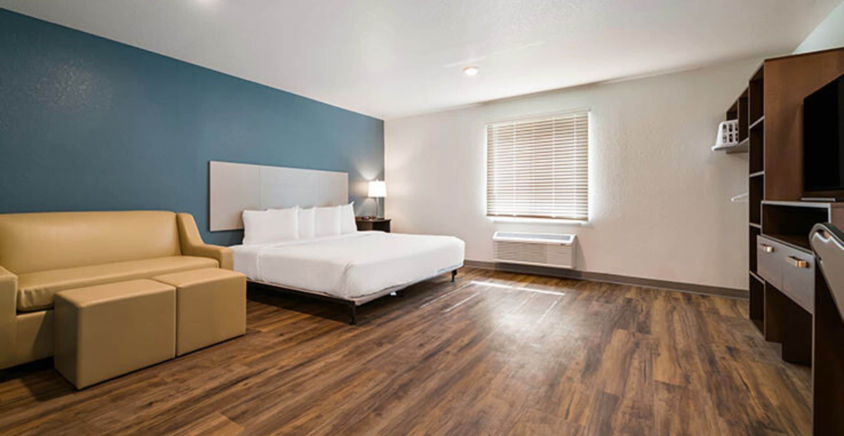 A hotel room with a bed, a sofa, a nightstand, a lamp, a window with blinds, and built-in shelves.