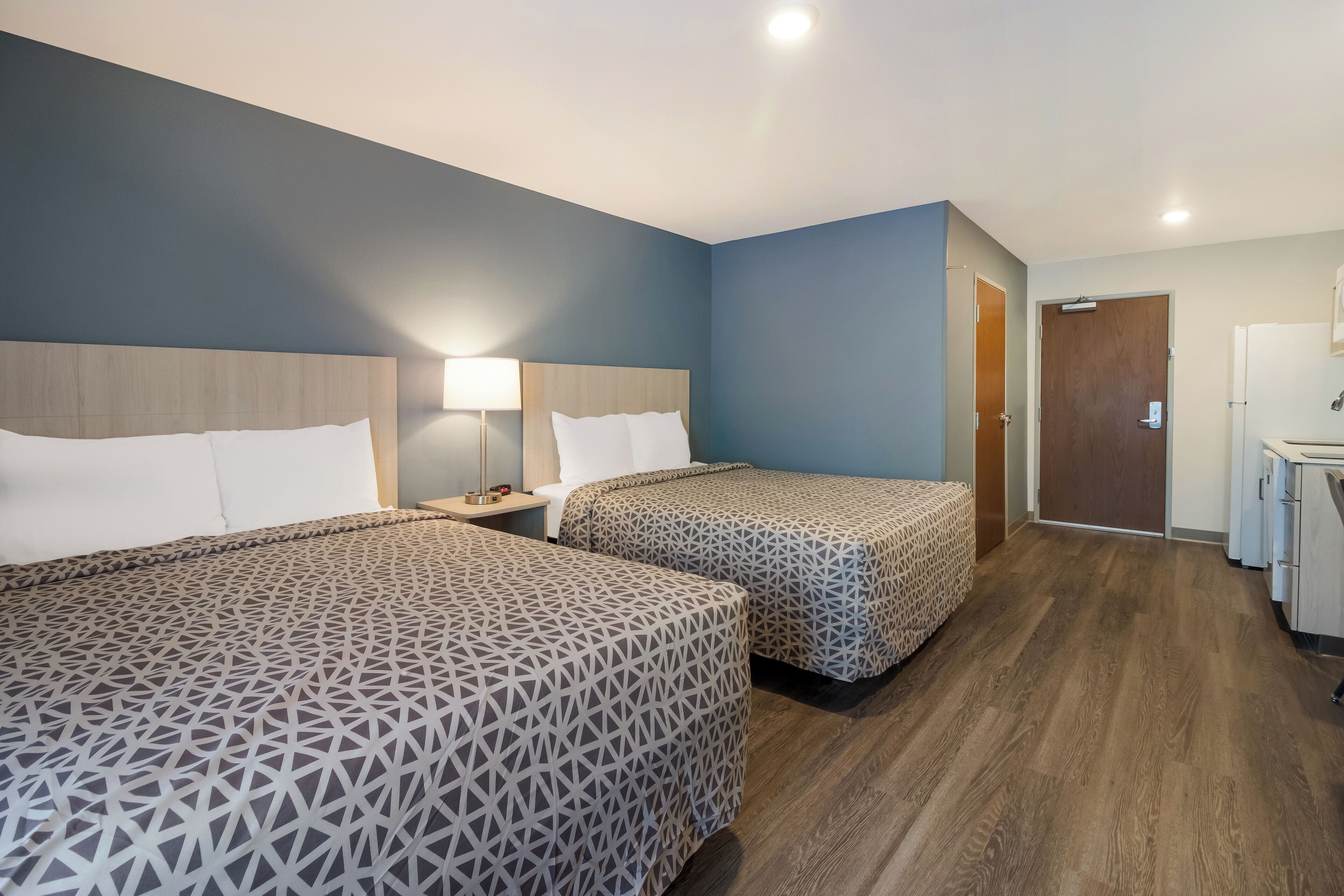 A hotel room with two beds, a nightstand with a lamp, a wooden headboard, a door, and a kitchenette.