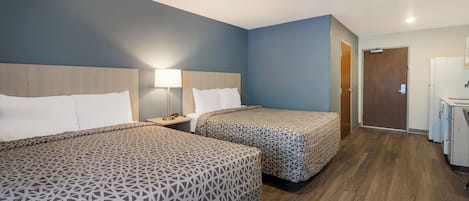 A hotel room with two beds, a nightstand with a lamp, a wooden headboard, a door, and a kitchenette.