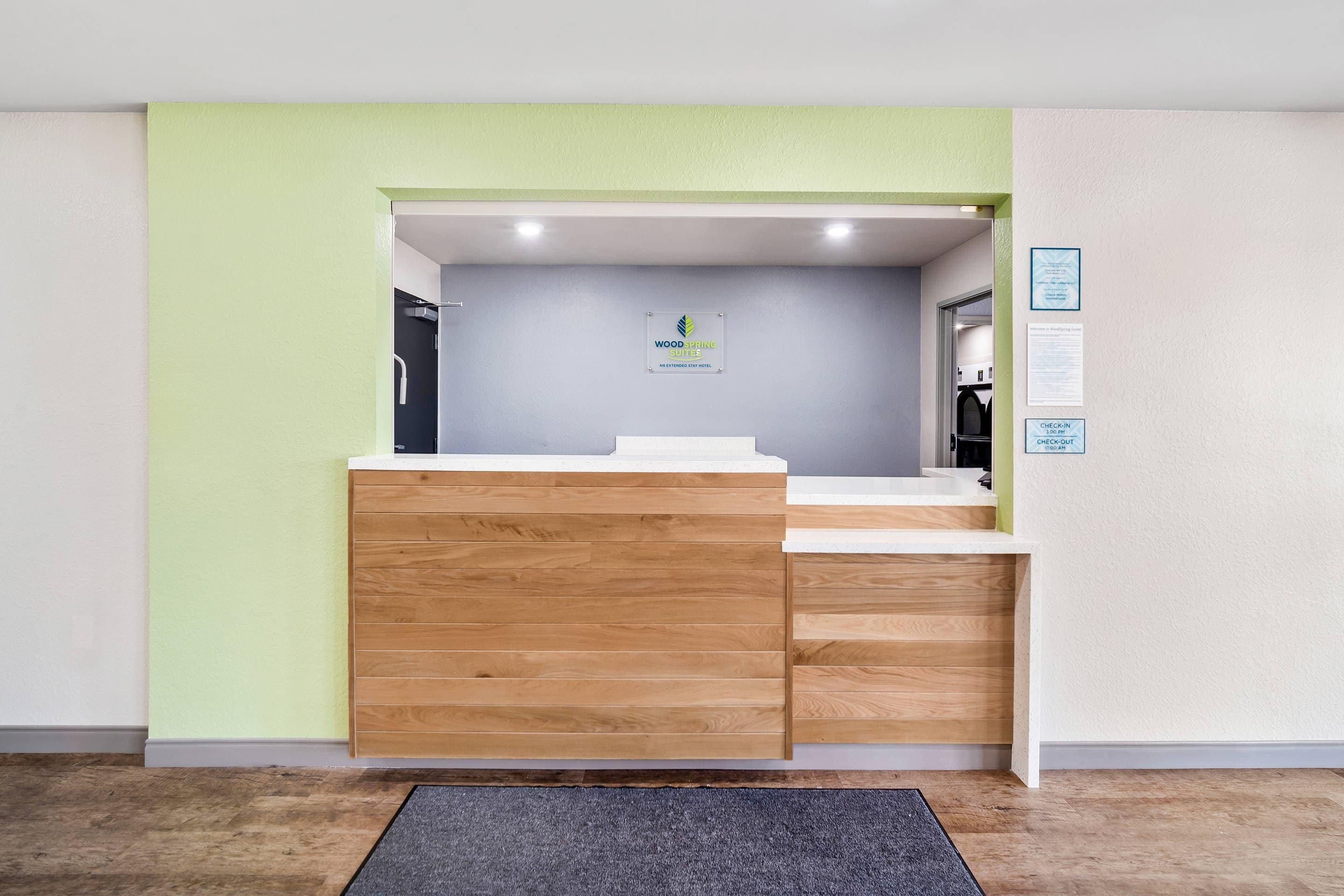 A reception desk with a wooden panel design, a check-in/out sign, and a sign indicating the hotel name 'WoodSpring Suites.'