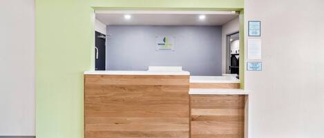 A reception desk with a wooden panel design, a check-in/out sign, and a sign indicating the hotel name 'WoodSpring Suites.'