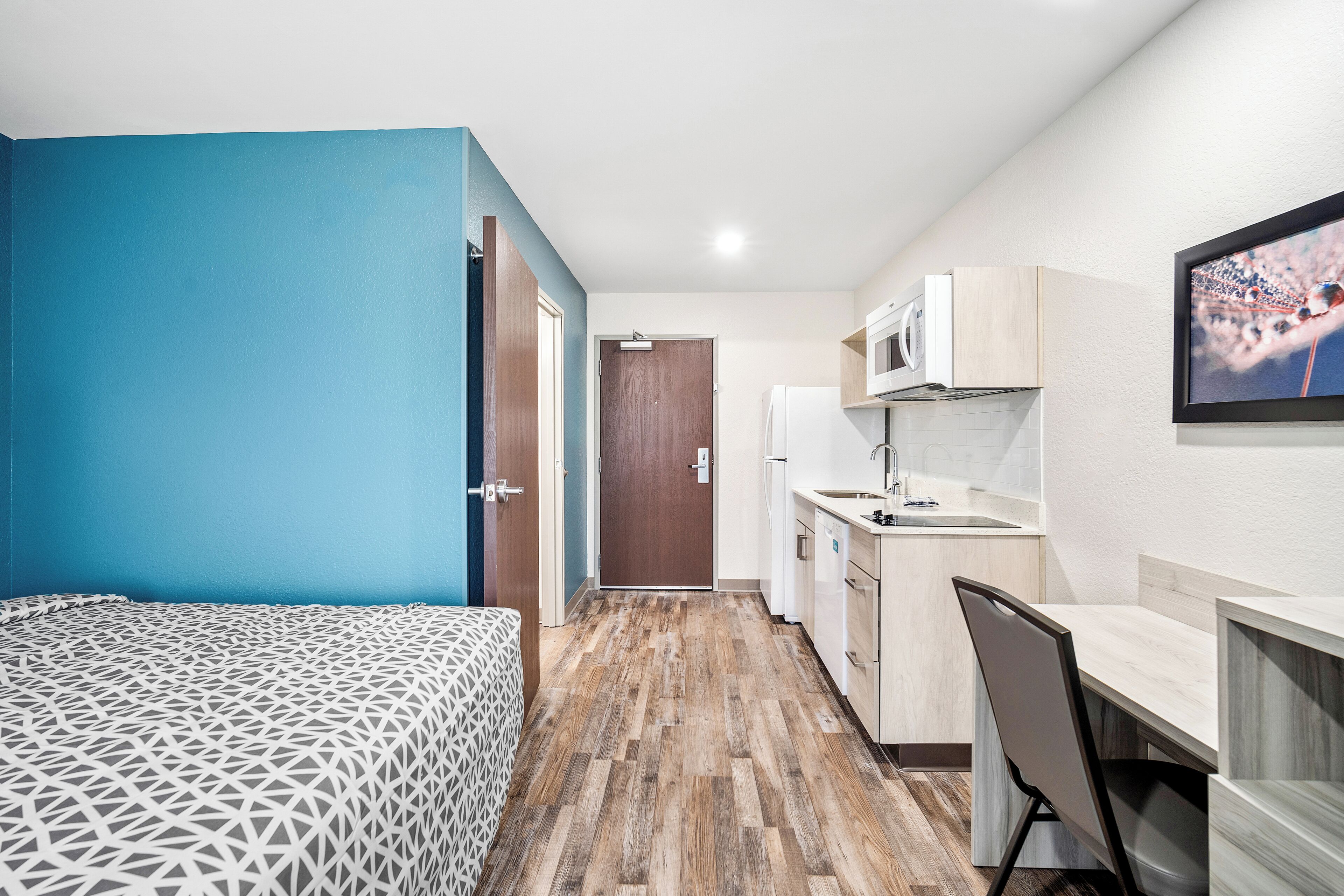 A compact room with a bed, kitchenette, and a door leading to another room.