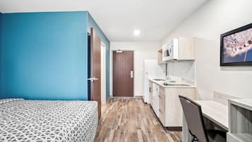 A compact room with a bed, kitchenette, and a door leading to another room.
