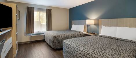 A hotel room with two beds, a TV, a desk with drawers, a window with curtains, and a lamp on the nightstand.
