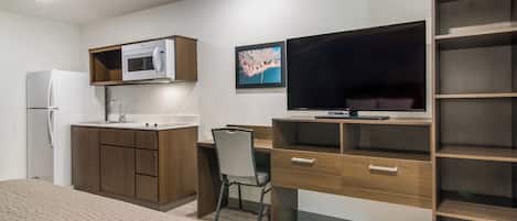 A compact hotel room with a kitchenette, a bed, a desk with a chair, a TV, and a bookshelf.