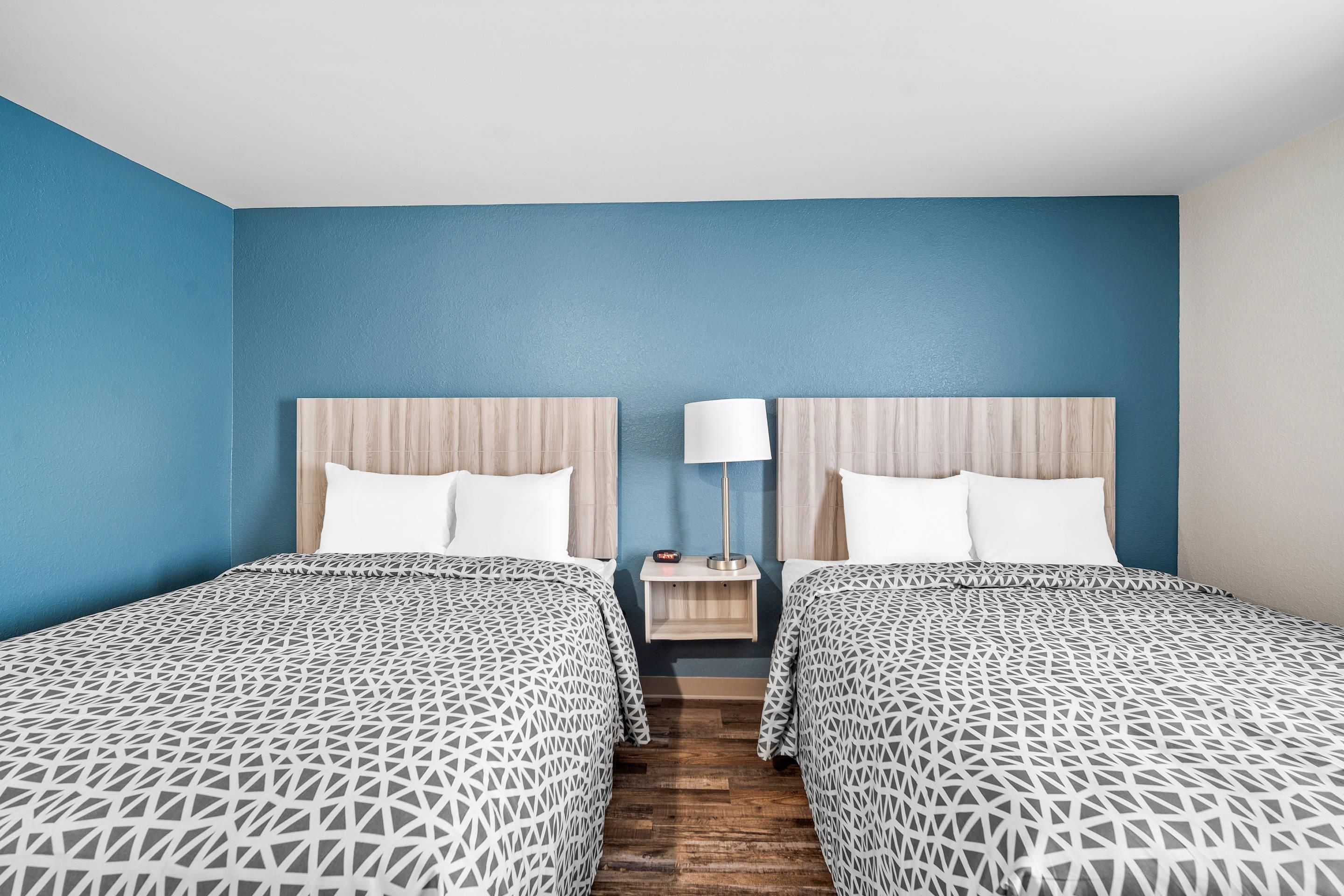 Habitación de hotel con dos camas, una mesita de noche, una lámpara y suelo de madera.