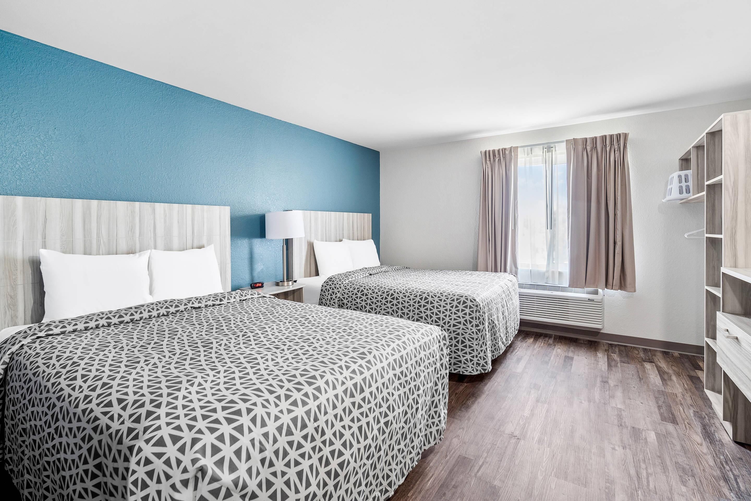 A hotel room with two beds, a nightstand, a lamp, a window with curtains, and a built-in air conditioning unit.