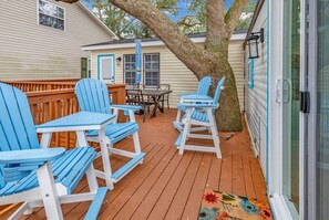 Eine Holzterrasse mit blauen Adirondack-Stühlen und einem Tisch, ein Baum und ein Haus im Hintergrund.